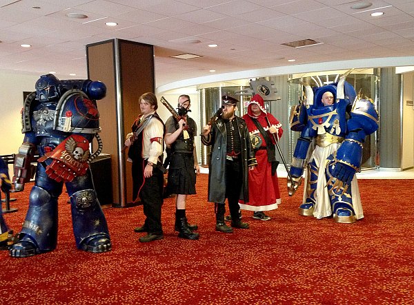 Шесть косплееров по вселенной Warhammer 40,000 стоят вместе в холле конвента: два крупных доспеха и четыре костюма из ткани