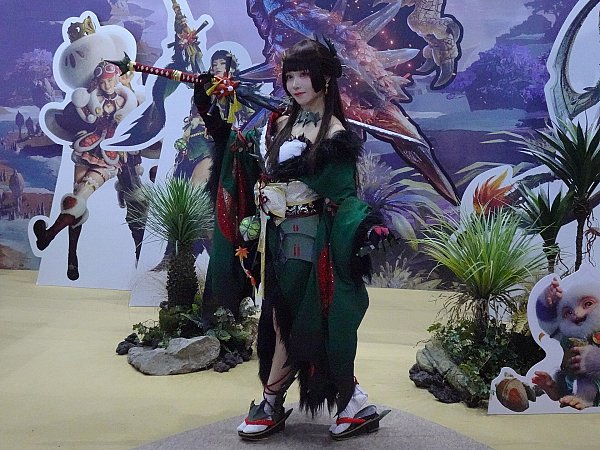 Косплеер в зелёном кимоно с длинным древковым оружием на фоне стенда Monster Hunter Outlanders